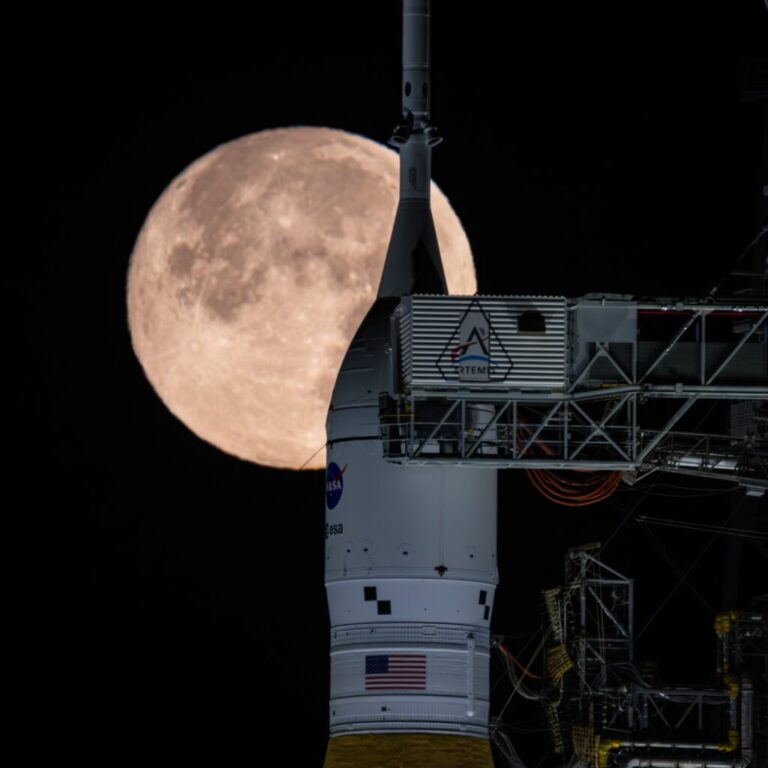 A large orange full moon rises behind a tall white rocket on a launch pad, with metal scaffolding around it at night.