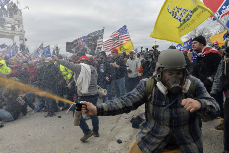 Rioters assault police protecting the U.S. Capitol on January 6, 2021. A man in the foreground is spraying an orange liquid. Crowds of people are packed together. Some are holding up flags, including the U.S. flag,