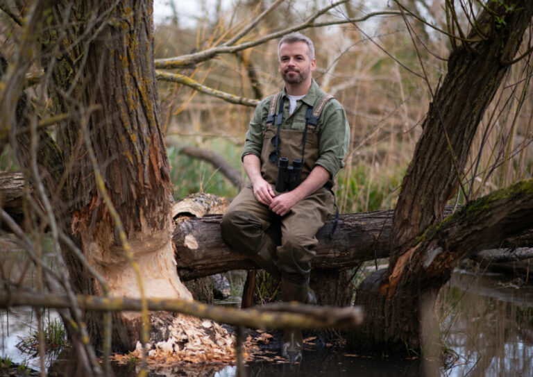 In this photo, Sean McCormack sits on a log in a marshy area near a tree whose trunk has been gnawed by beavers. He is wearing binoculars around his neck and overall-style waders.