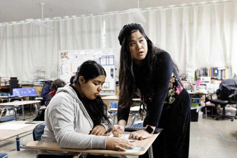 Mary Acebu, a Filipino woman wearing all black with a flowered vest and knitted beret, leans over a desk to help a student at her desk. The student has long brown hair in a ponytail and a gray sweatshirt.