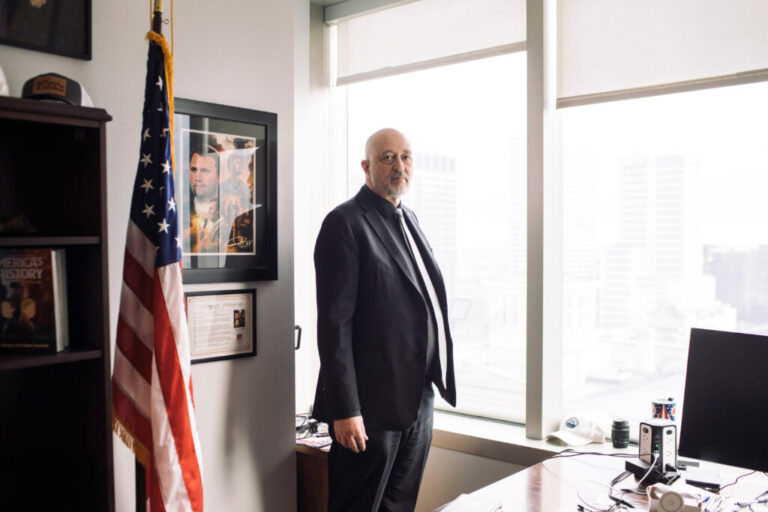 A man in a suit stands in an office next to a U.S. flag.