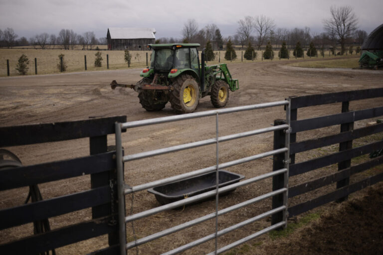A person drives a John Deere tractor down a dirt road at Hallstead Farms. A wooden fence with a metal gate is in the foreground. A wooden barn is in the background.