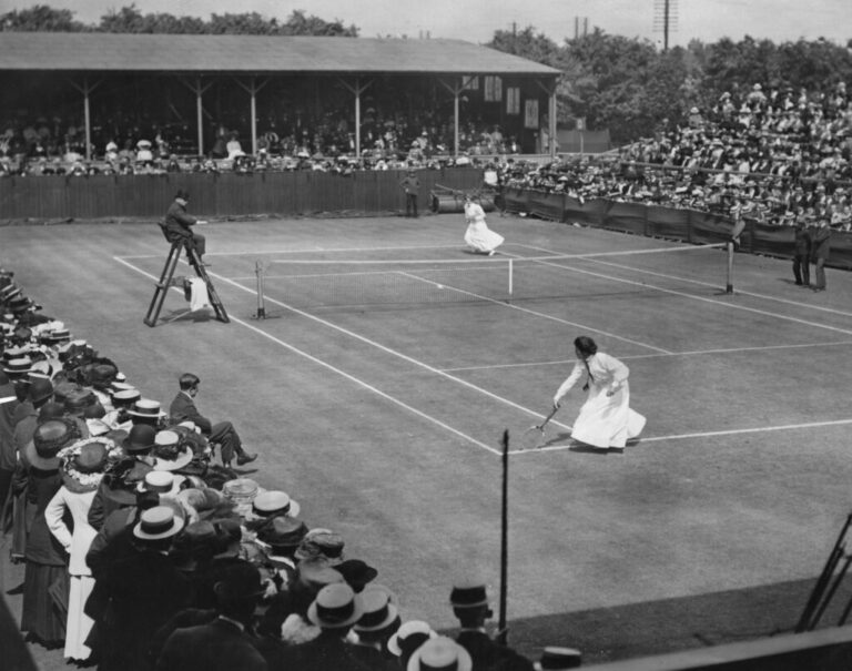 Edith Johnson and Dorothea Lambert Chambers, both wearing ankle-length white dresses, face off on a Wimbledon tennis court in 1910 in this black-and-white photograph.