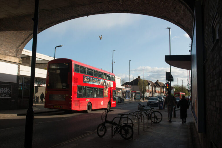 A red double-decker bus passes underneath a train bridge arch at Greenford Tube station.