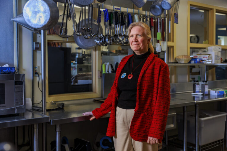 Mary Emerson is standing in the school's kitchen. Behind her is a shiny metal food preparation surface, and large spoons and ladles hang from a metal rod above.