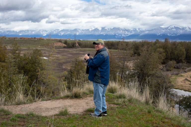 Mike Durglo Jr.  stands on a hillside above a river looking towards a mountain range. Durglo has devoted his life to preparing his home and his people for climate change. As the Confederated Salish and Kootenai Tribes climate change coordinator he wrote one of the first tribal climate action plans in the country over 15 years ago.