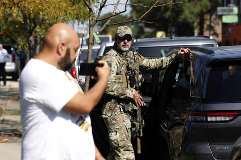 A man hold his phone up next to a Border Patrol agent dressed in fatigues and resting one arm on a vehicle parked at the curb.