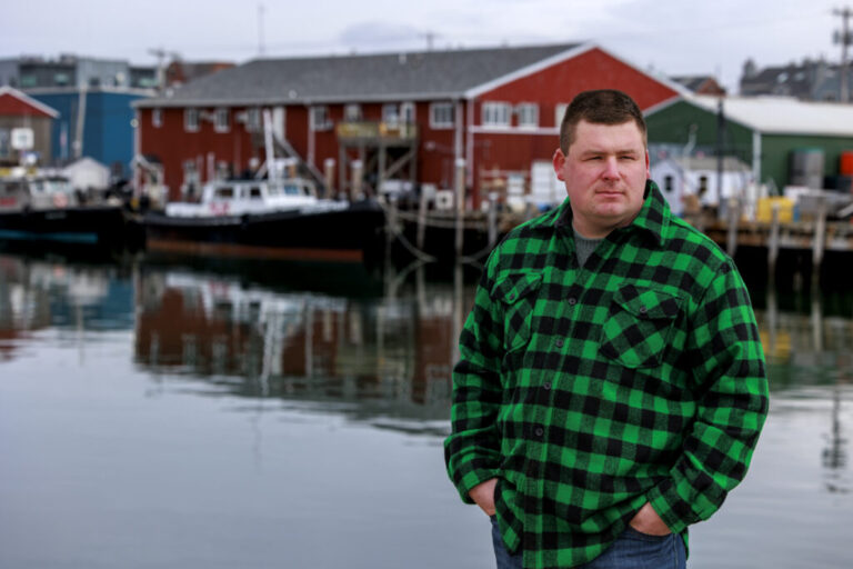 Fisherman Devyn Campbell stands near the water outside the Portland Fish Exchange. Boats and a marina are behind him.