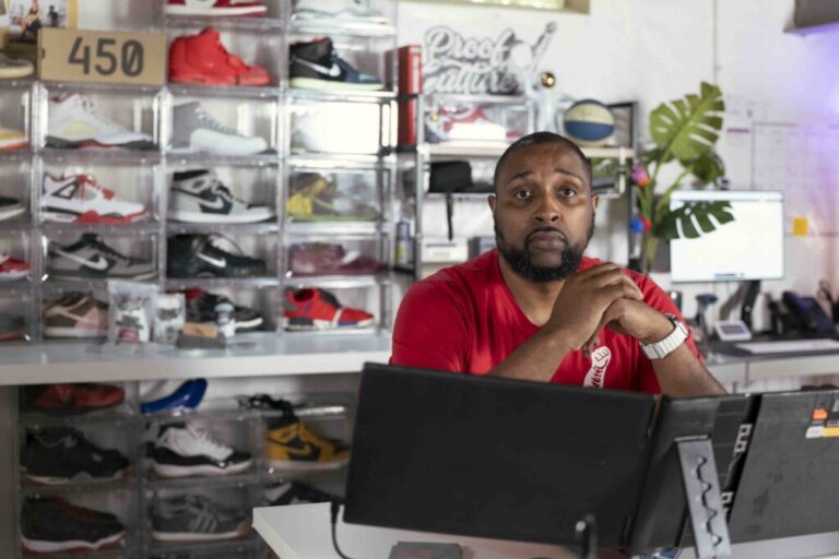 Richard Brown runs Proof Culture, a sneaker accessory company, out of his Ohio home. He is Black and has a beard. He is seated behind a computer monitor and there are sneakers in plastic display cases behind him.