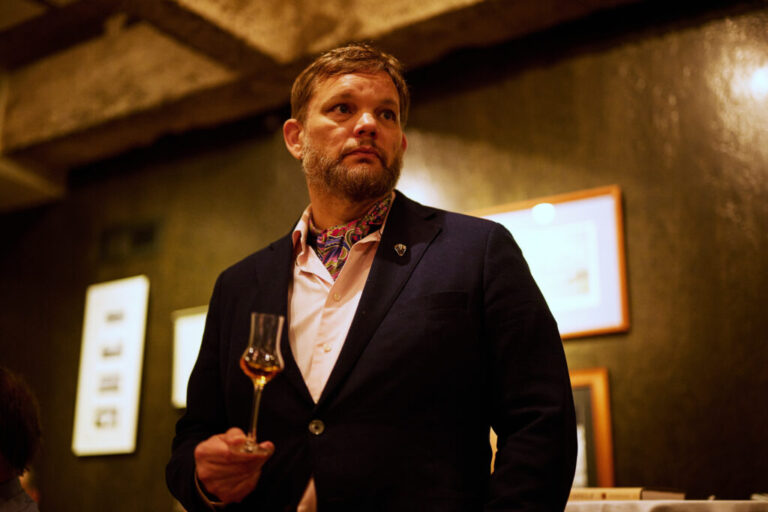 Iraq war combat veteran Fred Minnick holds a small glass of bourbon during a tasting dinner at Cordelia, a restaurant in Cleveland's historic district. He is wearing a blazer and a patterned cravat around his neck tucked into his button down shirt. He has blond hair and facial hair.