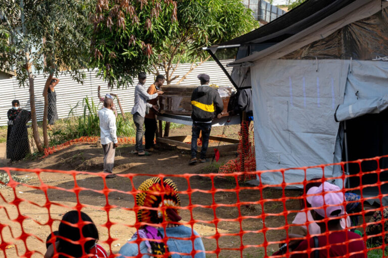 An outside view of the morgue at the Ebola treatment center of the Evangelical Medical Center in Bunia, Ituri province, Democratic Republic of Congo, on June 18, 2026. The morgue is covered in tarp on the outside. Next to the morgue, three men are handling a brown coffin that's on a table.