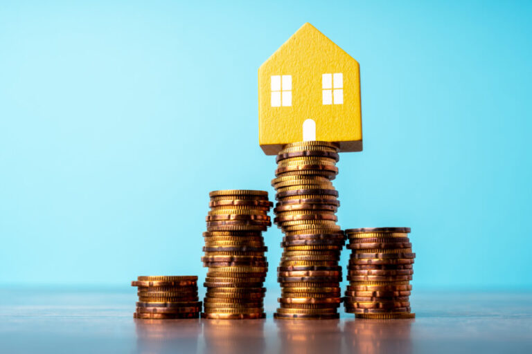 This photo shows four stacks of coins of varying heights standing in a row. At the top of the tallest stack, the second stack from the right, sits a small model of a yellow house with a front door and two windows. The background of the photo is light blue.