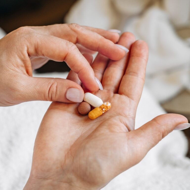 A hand with nude-painted nails lifts a single pill from a group of three in the other hand's palm.