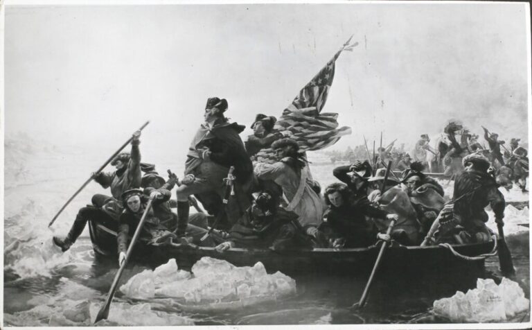 This black-and-white image shows George Washington crossing the Delaware River during the American Revolutionary War in 1776. He's standing in a rowboat as men around him paddle the boat through the river, which has large chunks of ice floating in it. Two men behind him in the boat hold a partially unfurled American flag.