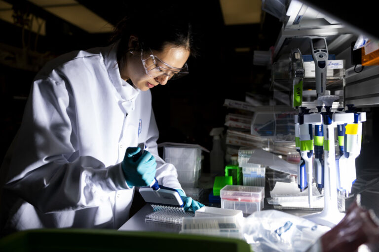 In this photo, a researcher works in a lab at the Moderna headquarters in Cambridge, Massachusetts. She's wearing a white lab coat, safety goggles and blue gloves and is working with a set of pipettes.