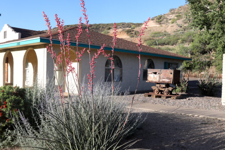 An old mine cart is parked outside the Gila County Historical Museum in Globe, Arizona. The small building is in a desert landscape.