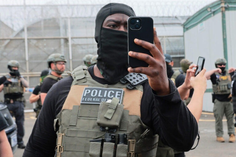An Immigration and Customs Enforcement officer, wearing tactical face covering and vest, holds up a phone to shoot video.