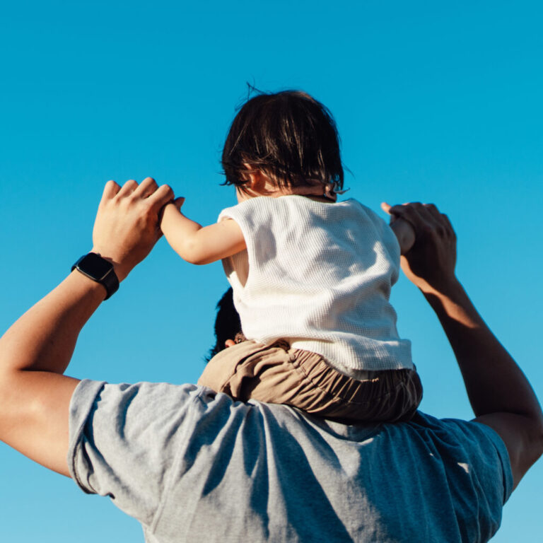 Rear view of toddler on father's shoulders against blue sky.