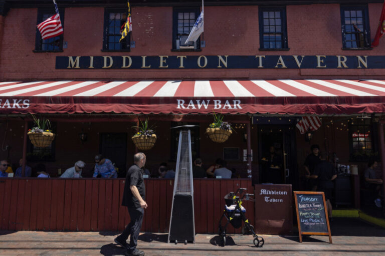 A man dressed in all black walks in front of the Middleton Tavern, which is housed in red brick building and has a maroon and white awning covering a few patrons seated outside.