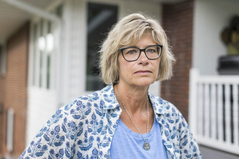 Maria Ruhtenberg has chin-length blond hair and she is wearing glasses, a blue and white patterned shirt over a light blue T-shirt. She is standing in front of a house that has brick and white siding.