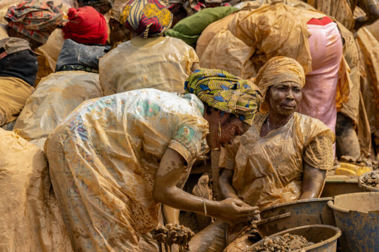 Women wearing plastic protective garments sift gold-bearing sediments at the Maidede mine in Mongbwalu, Ituri province, Democratic Republic of Congo, on June 16, 2026. Much of their clothing and bodies are covered in light brown sediment.