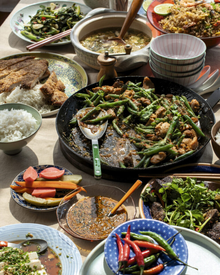 This photo shows a green bean and chicken stir-fry, with sauce and seasonings, in a black pan. It is surrounded by other dishes that include vegetables and rice.