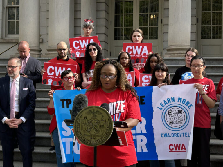 Valerus speaks at a LanguageLine press conference in front of New York City Hall in April. All the workers and organizers are wearing red T-shirts. Valerus has long frizzy hair and wears glasses.
