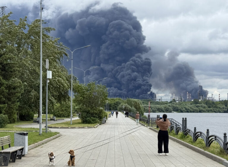People walk in a park as black smoke rises from the area of the Russian oil producer Gazprom Neft's Moscow oil refinery