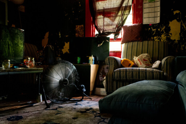 This photo shows a room with a floor fan on a rug, an upholstered armchair, an end table and a coffee table. A sheer curtain covers the window, darkening the room.