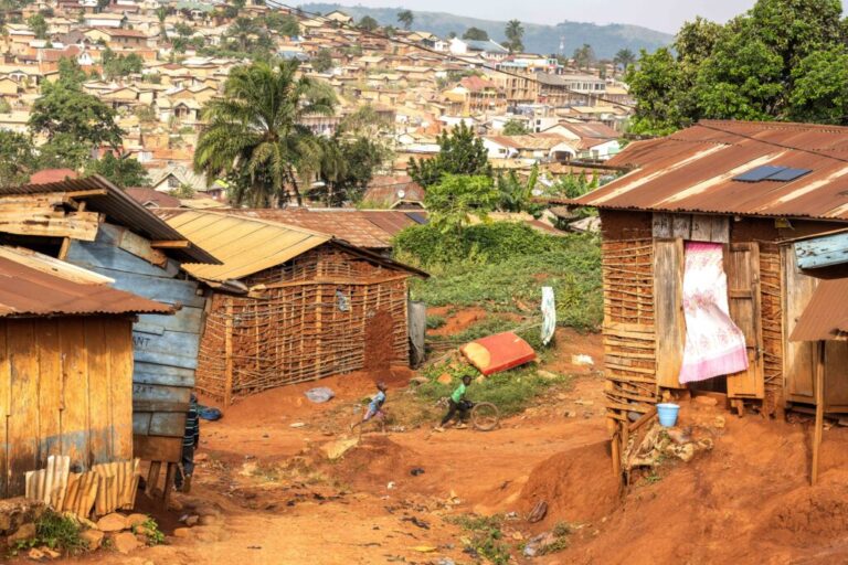 A general view of the Shuni neighborhood in Mongbwalu, Democratic Republic of Congo, on June 16, 2026. Two boys play, with one boy rolling a tire forward and the other boy following behind. The neighborhood has homes built from wood and corrugated metal.