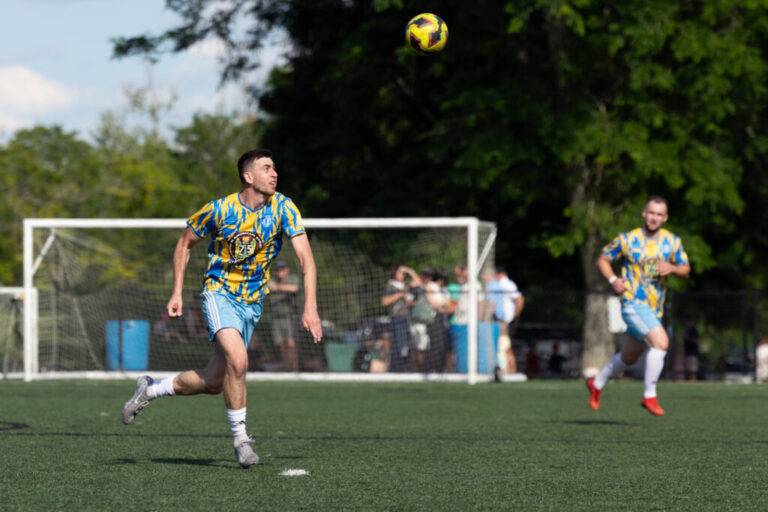Standing on a green soccer field and wearing a yellow and light blue soccer uniform, Oleksandr “Sasha” Ostapchuk sets himself up to head a ball that's in the air while playing for a team of Ukrainian immigrants against a club with roots in Belarus at Tryzub Ukrainian American Sport Center in North Wales, Pennsylvania, on June 21.