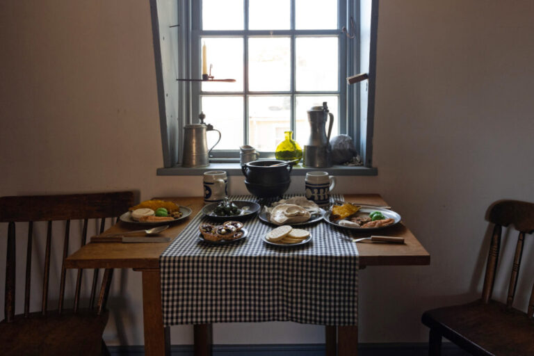 A table setting depicting typical of working-class people meal in 18th-century Annapolis is displayed at Hogshead Trades Museum in Annapolis. The table has flat breads, small pies, large ceramic mugs and is placed under a window.