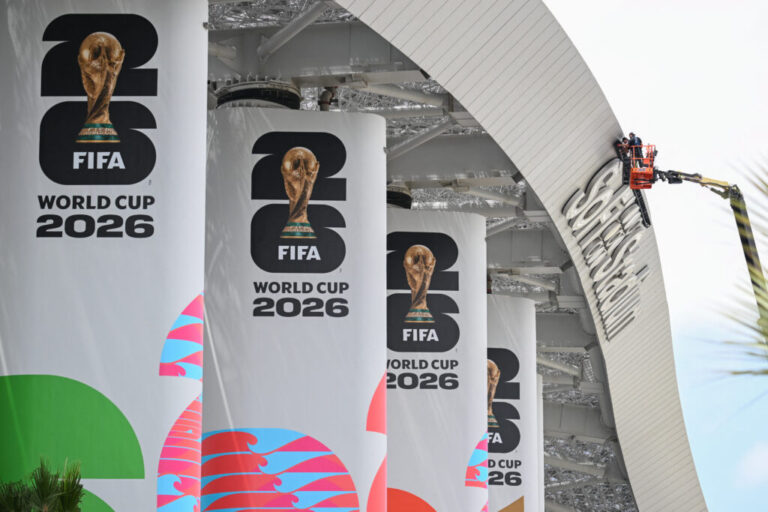 Workers work from a crane at Los Angeles Stadium on June 7, ahead of the 2026 FIFA World Cup. The workers are standing in a metal basket at the end of the crane's long arm and are doing work to the top rim of the stadium.