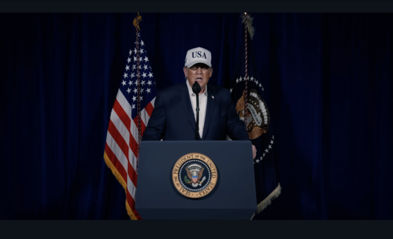 In this screenshot from a February 28 video, President Trump stands behind a lectern to discuss strikes on Iran. Speaking into a microphone, he's wearing a white baseball cap that says