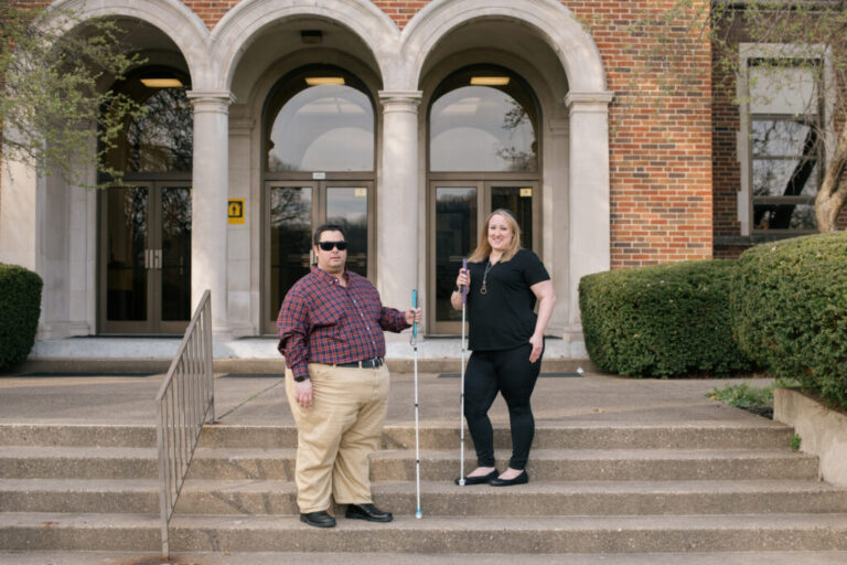 ALT TEXT: A 34 year old white man with short, black hair, dark glasses, and a plaid shirt stands to the left. A 43 year old woman with shoulder length blonde hair and an all black outfit, stands to the right. They’re both holding canes on stairs in front of a brick building.