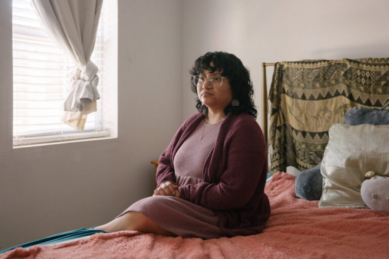 Lourdes Monje, a young person with curly dark hair and glasses, sits on a bed next to a window. They are wearing a mauve colored dress and sweater.