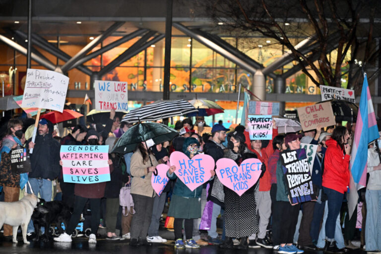 A large gathering of people are shown, most carrying homemade signs, in front of a hospital building. It is raining and people have umbrellas open.