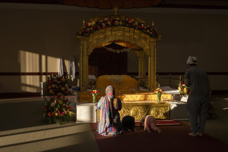 A family bows on their knees, touching their heads to the ground in worship before the Guru Granth Sahib, the eternal Guru of the Sikh faith, at the Guru Nanak Society of Greater Cincinnati. In Sikhism, spiritual guidance once came from human Gurus, beginning with Guru Nanak and continuing through nine successive Gurus. After the tenth Guru, Guru Gobind Singh, the lineage of human Gurus ended, and the Guru Granth Sahib—the holy scripture was declared the eternal Guru, embodying their wisdom, teachings, and spiritual presence. The text rests on a raised throne and is treated with the same respect as a living person. it is ceremonially awakened each morning, offered food, and ceremoniously laid to rest at night. Devotees gather to sing hymns and offer prayers, practicing devotion, humility, and service. These traditions preserve a living connection to the Gurus’ teachings and pass cultural and spiritual values to future generations, anchoring the Sikh identity even while living in the America.