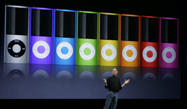 Apple's then-CEO, Steve Jobs, stands on a stage to introduce new colors for the iPod Nano in September 2008. Behind him is an enlarged image of a row of iPod Nanos in seven rainbow colors and gray.