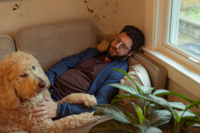 Benjamin Stein-Lobovits, a young man who has dark curly hair, a beard and glasses, lays on the couch petting his dog, Cow, a large, beige poodle mix.
