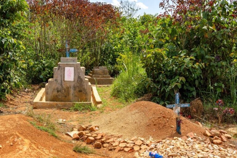 A general view of a cemetery in Mongbwalu, Ituri province, Democratic Republic of Congo, on June 17, 2026. Three graves can be seen, one of which is marked by a mound of dirt and a blue cross staked into the mound. Lush green foliage rises in the background.