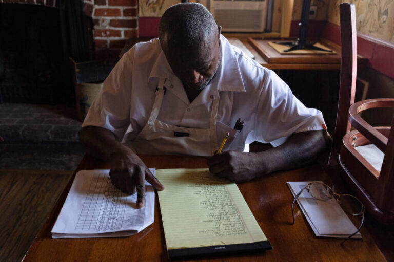 Executive chef Arthur Gross, a Black man, sits in an upstairs dining area to work on his handwritten ingredient order. He is wearing a white button up short sleeve shirt with a white apron on top.