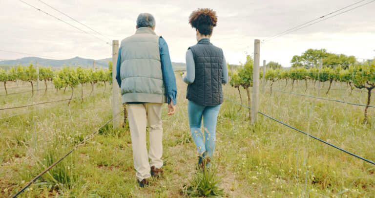 Photographed from the rear, a man and a woman walk in a grassy area on a farm, with slopes rising up in the background.