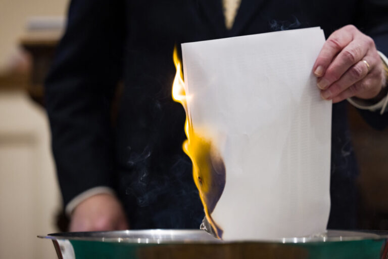 A flame is seen on one side of a burning piece of white paper; the Rev. John Jackman holds it carefully on the other side.