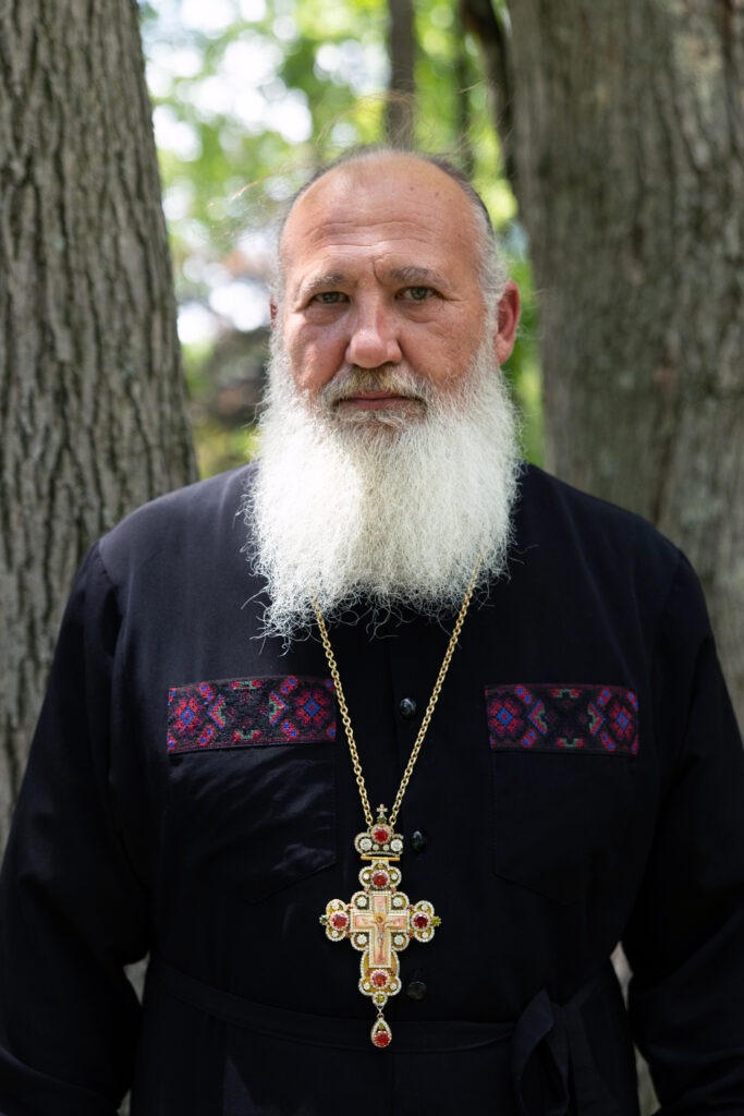 Father Taras Naumenko poses for a photo on June 21, 2026. He is wearing a cross around his neck and is photographed from about the waist up. He's standing outdoors, and two tree trunks rise behind him.