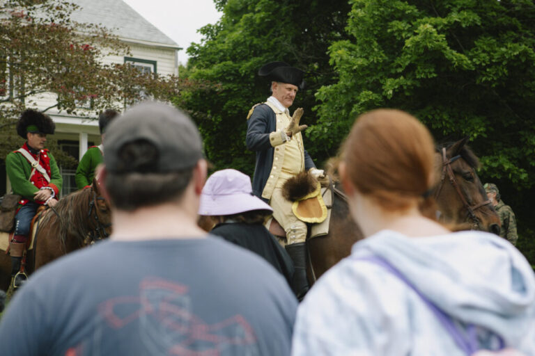 A reenactor dressed as George Washington, wearing a  tricorn hat, waves to the crowd from atop his horse.
