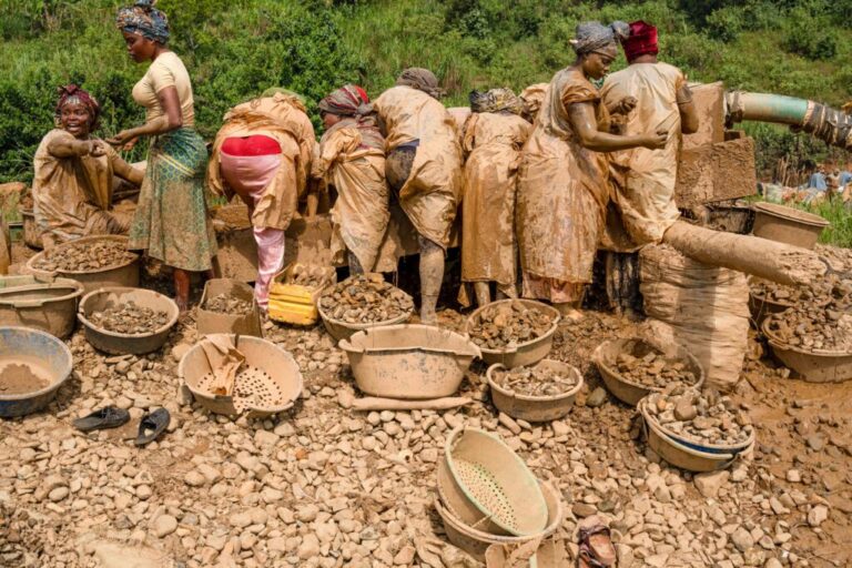 Women wearing plastic protective garments sift gold-bearing sediments at the Maidede mine, in Mongbwalu, Ituri province, Democratic Republic of Congo, on June 16, 2026. Much of their garments and bodies are covered in light brown sediment. Near them on the ground are basins filled with rocks.