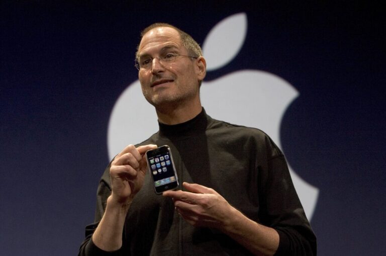 In this 2007 photo, Steve Jobs, then Apple's CEO, holds an iPhone during the smartphone's unveiling. Jobs is wearing a black turtleneck shirt, and he's standing in front of a black backdrop that's displaying the Apple logo in white.
