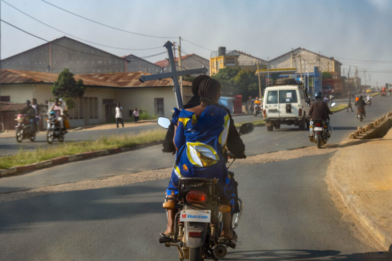 Photographed with her back to the camera, a woman carrying a funeral cross rides on the back of a motorcycle taxi through the streets of Bunia, Ituri province, Democratic Republic of Congo, on June 18, 2026. Other motorcycles are traveling on the road in both directions.