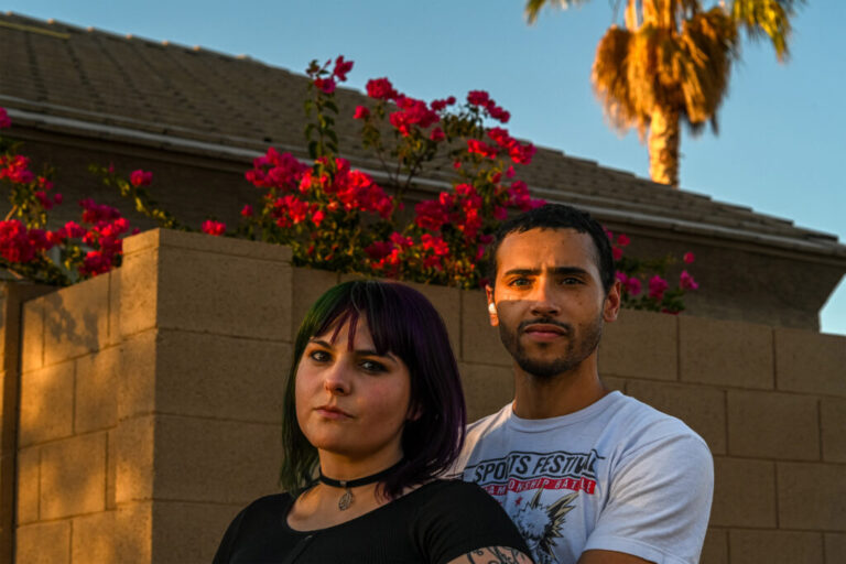 Samantha and Ariane Buck are a young couple standing in front of a house with a high-walled garden. Bright red flowers peek over the wall above their heads.